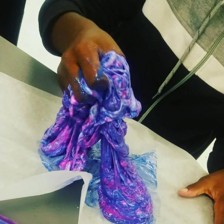 A camper’s hand lifts a stretch of purple, pink and blue slime off a sheet of wax paper on a table.